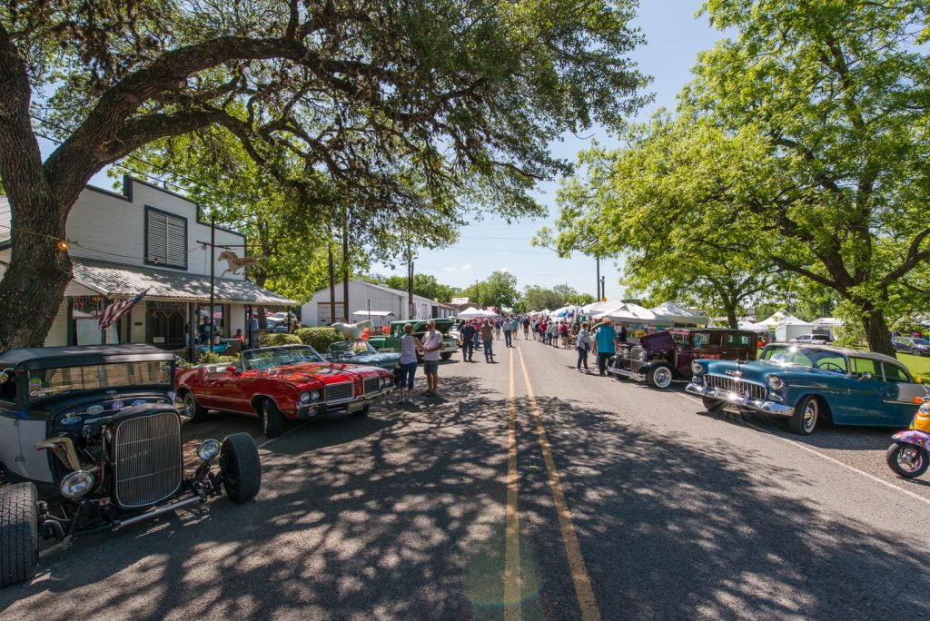 A street in Downtown Burton. Both sides of the street are lined with classic cars. In the back there are vendor tents set up and groups of people are walking around.