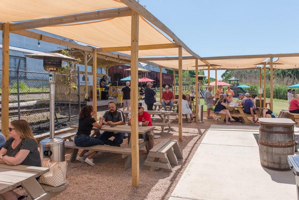 groups of people sitting at picnic tables under shade structures, talking and enjoying live music outside of Brazos Valley Brewery