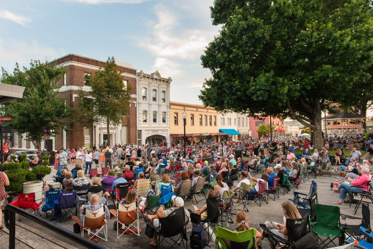 large audience of people sitting on two intersecting streets of downtown Brenham for Hot Nights Cool Tunes, Saturdays in July. Live band performing in the far left of the image.