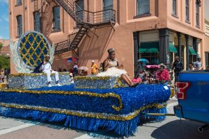 Juneteenth Jr Queen and King sit on top of a blue, silver, and gold float, waving to attendees.
