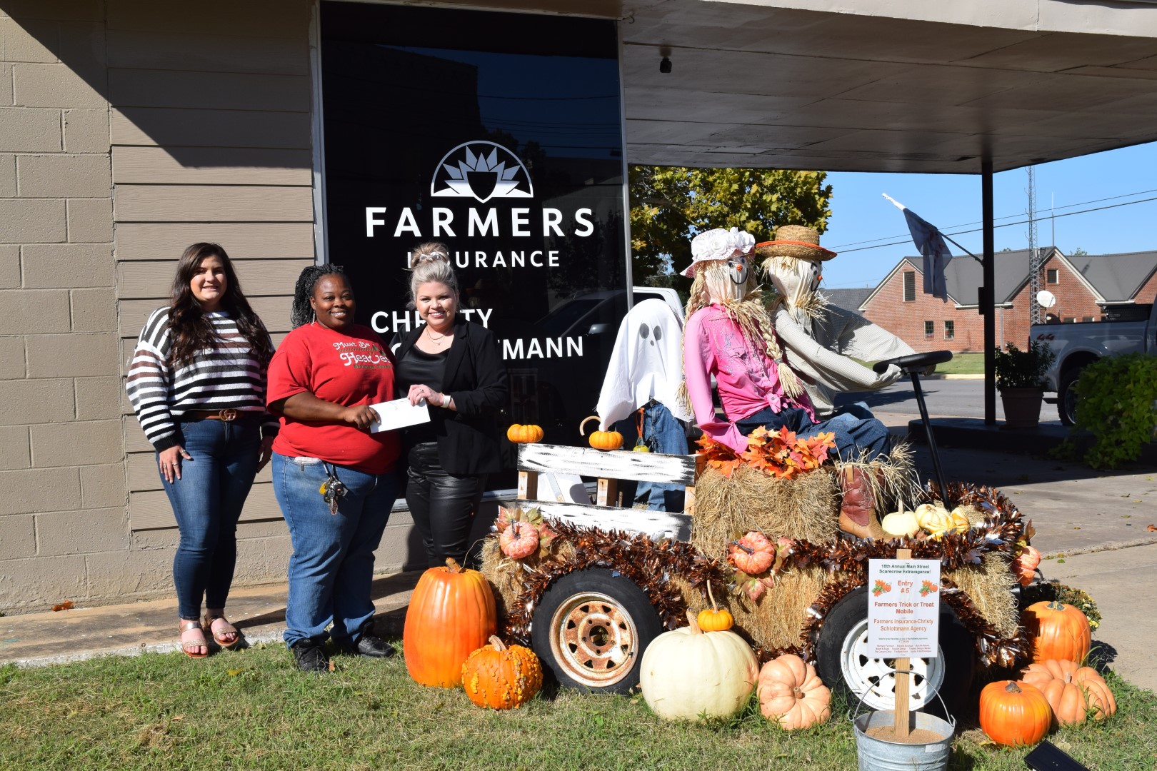 Three women in front of farmers insurance standing next to a truck made out of hay with two scarecrows sitting in the front and one in the bed of the "truck" in a ghost costume.