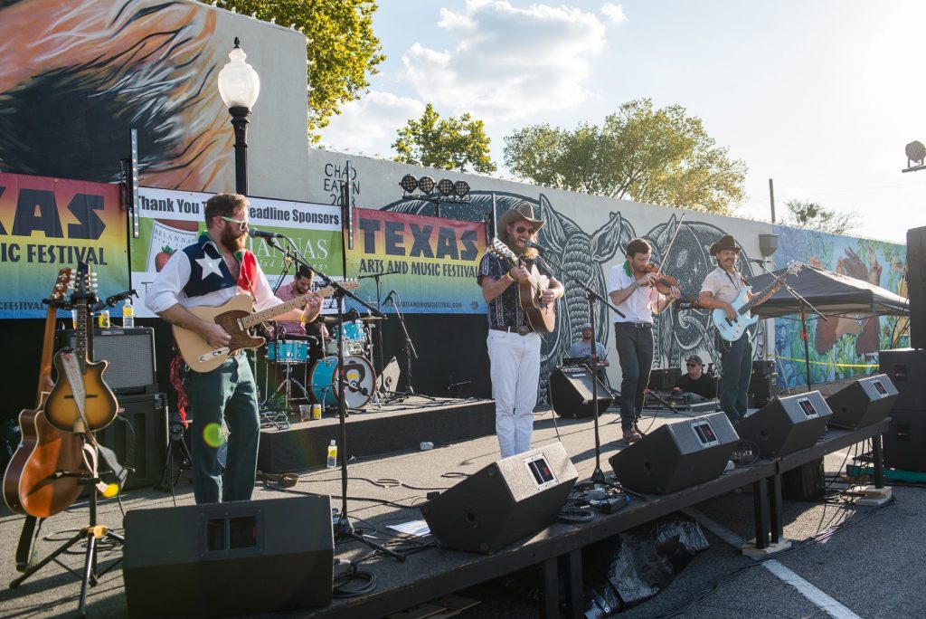 Band performing on stage in front of mural in Commerce Street parking lot during Texas Arts & Music Festival.