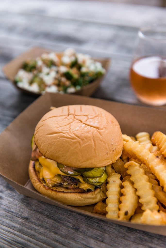 Burger and fries in a paper container with blurred out food and wine in the background. From Country Sunshine Food Truck.