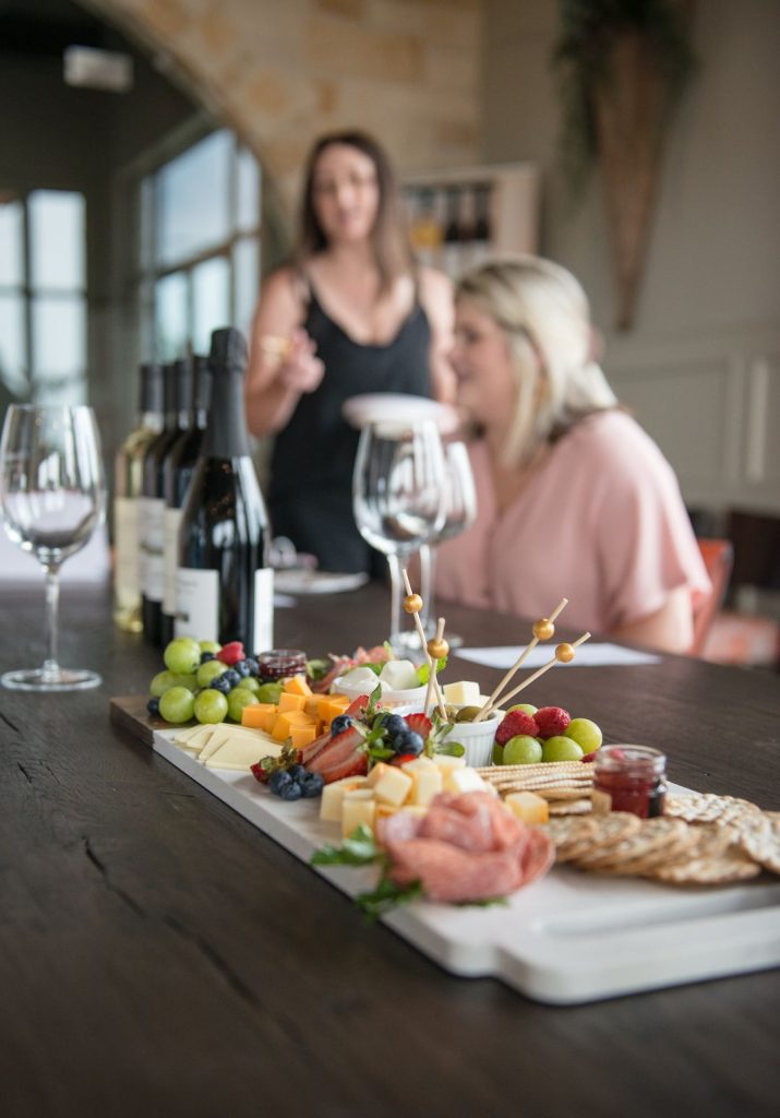 Charcuterie board with bottles of wine and glasses in the background, along with two ladies blurred out.