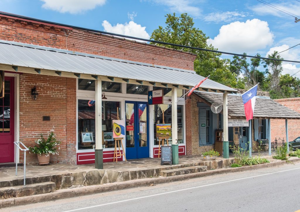 "Texas Artisans" wooden sign hangs on an awning covered sidewalk abutting the storefront. a painting of a landscape and a still life avocado sit on easels next to the glass and wood frame doors.