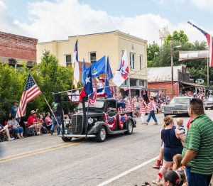 a black classic car in a parade with many flags and red, white, and blue, for independence day in Chappell Hill