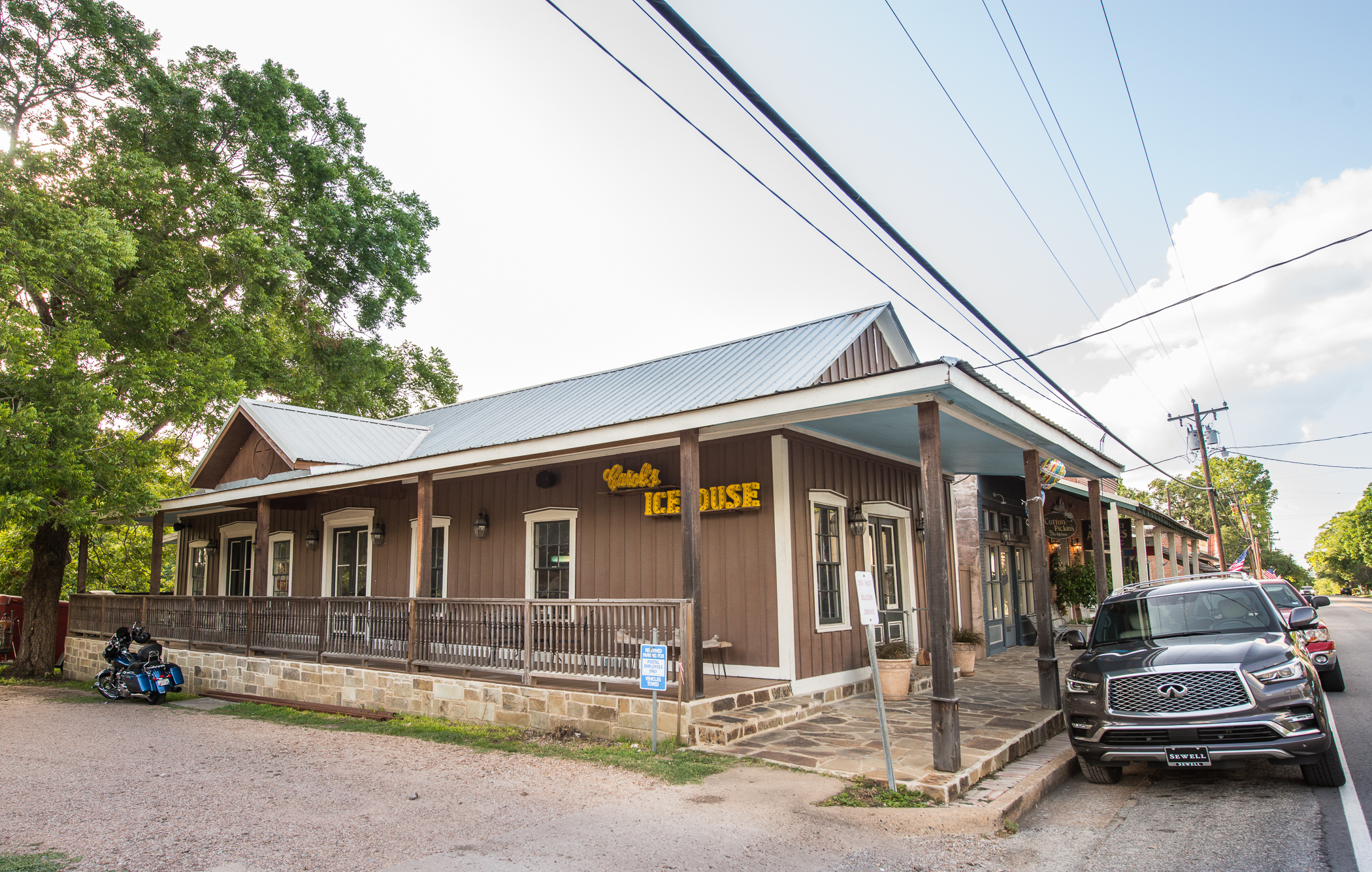 Exterior view of Carol’s Ice House in Chappell Hill, Texas, a rustic brown wooden building with a tin roof and stone foundation. A yellow sign reading “Carol’s Ice House” hangs above the porch, where wooden posts support a covered walkway. A motorcycle is parked on the gravel beside the building, and cars line the street under overhead power lines on a sunny day.