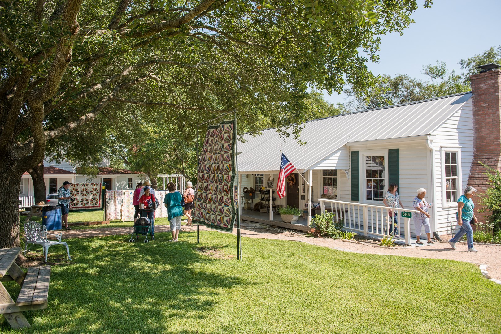 Quilts are hung up outside and people are walking around looking at them. White building with American flag is in the back.