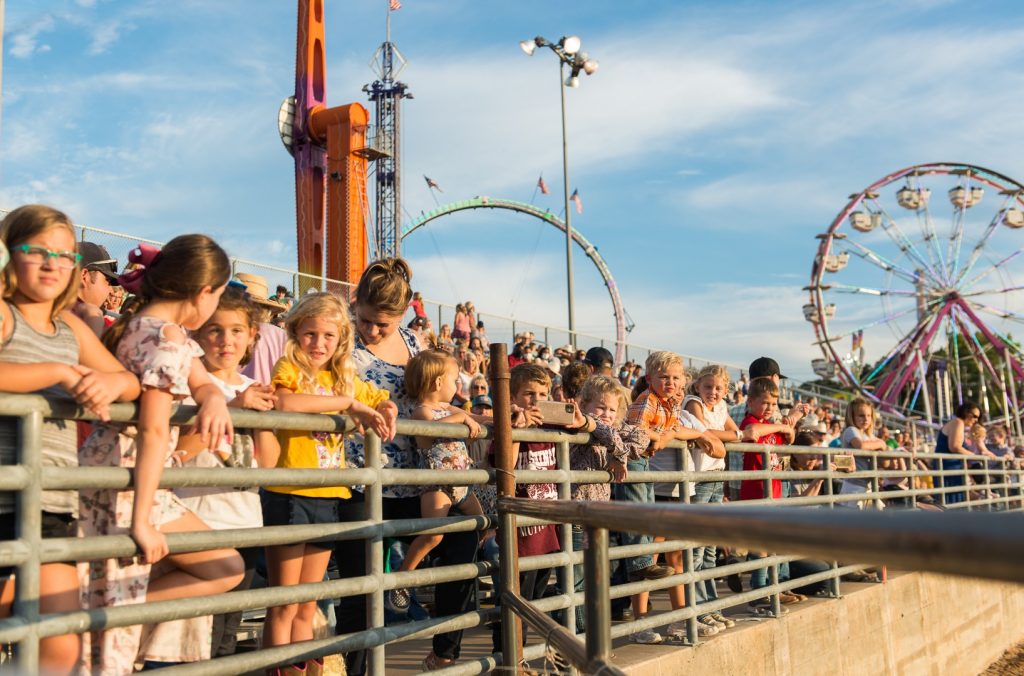 Crowds of people, especially children, leaning against a railing to watch the rodeo. Ferris wheel and other carnival rides are in the background. Washington County Fair.