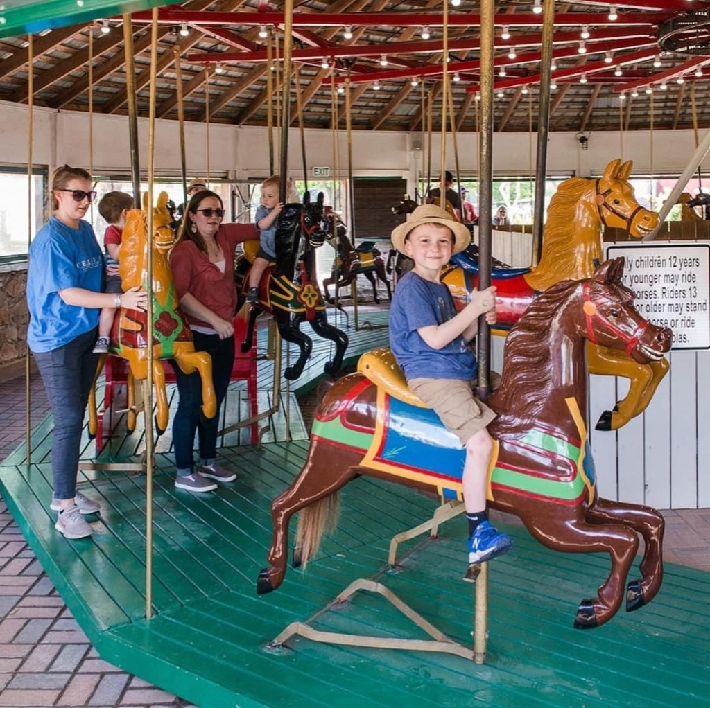 child riding antique carousel ride with other children riding it in the background. Adults standing in back.
