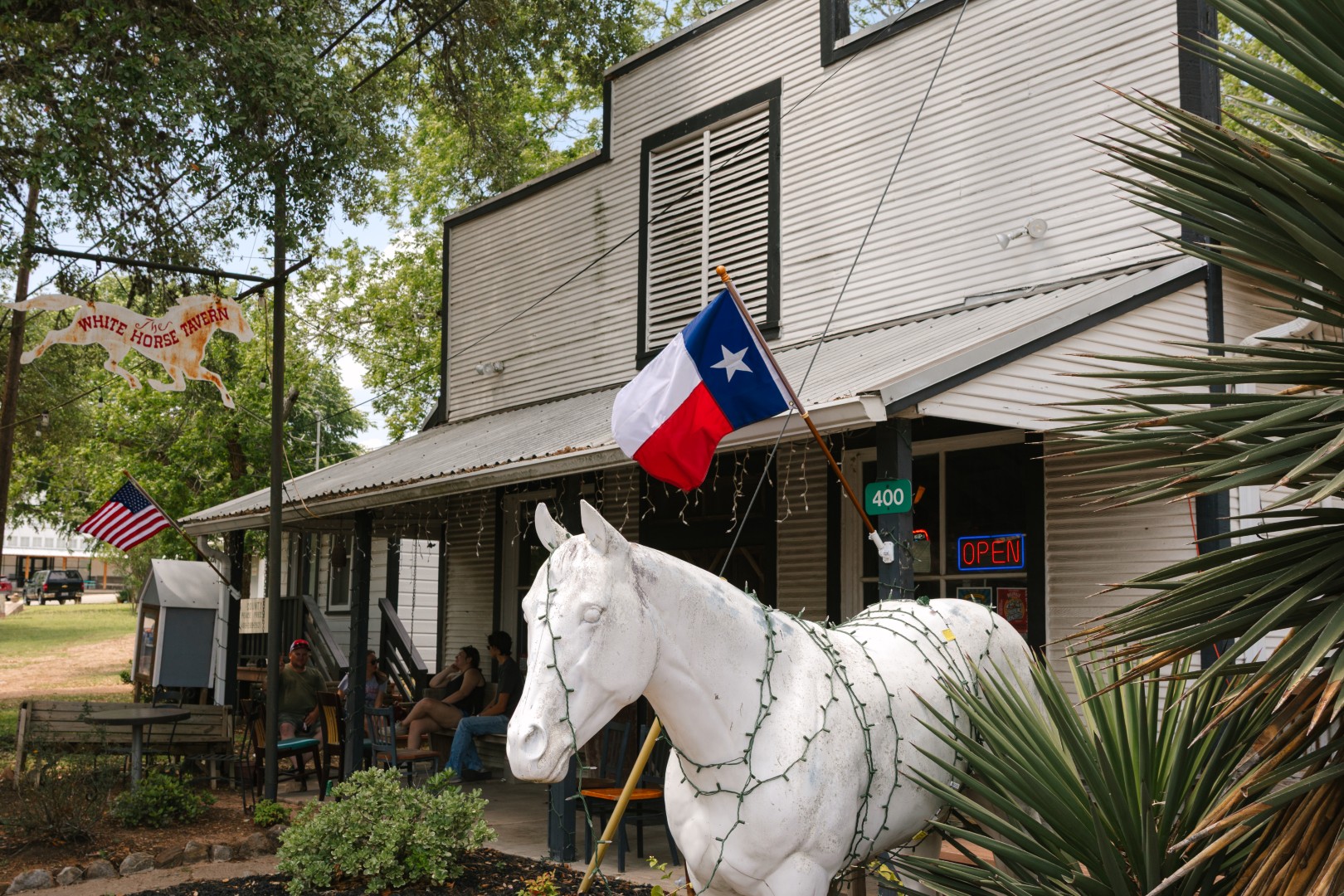 Exterior of The White Horse Tavern in Burton, Texas, with a rustic metal-sided building and a front porch where people are seated. A large white horse statue decorated with string lights stands out front beneath a Texas flag. A hanging sign with a galloping horse reads “The White Horse Tavern,” and a neon “OPEN” sign glows near the entrance.
