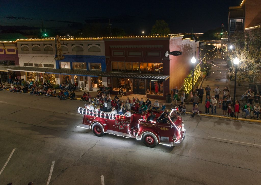 Christmas parade firetruck going through the streets of Downtown Brenham. Groups of people are on the sidewalk watching.