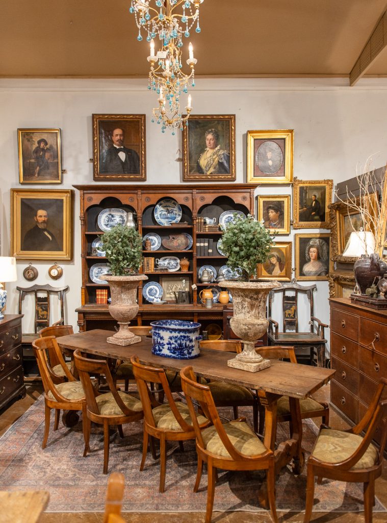 The inside of an antique store. There is a dinning table in the center with 10 chairs around it. A chandelier with blue beads is hanging above it. On the wall behind the table is old painted portraits of both men and women.