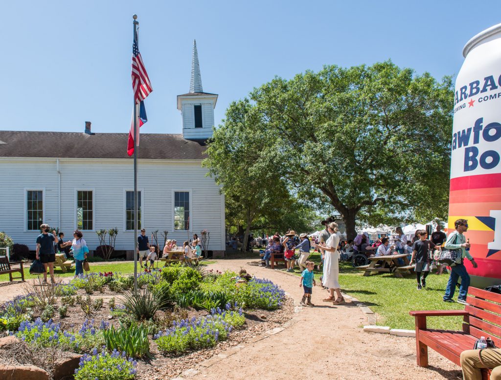 Many children and adults outside for a festival in front of a white wooden chapel. To the left there are white tents and a large inflatable of a Karbach beer in the Houston Astros colors. There is an American and Texas flag on a flag pole in the center with some patches of bluebonnets surrounding it.