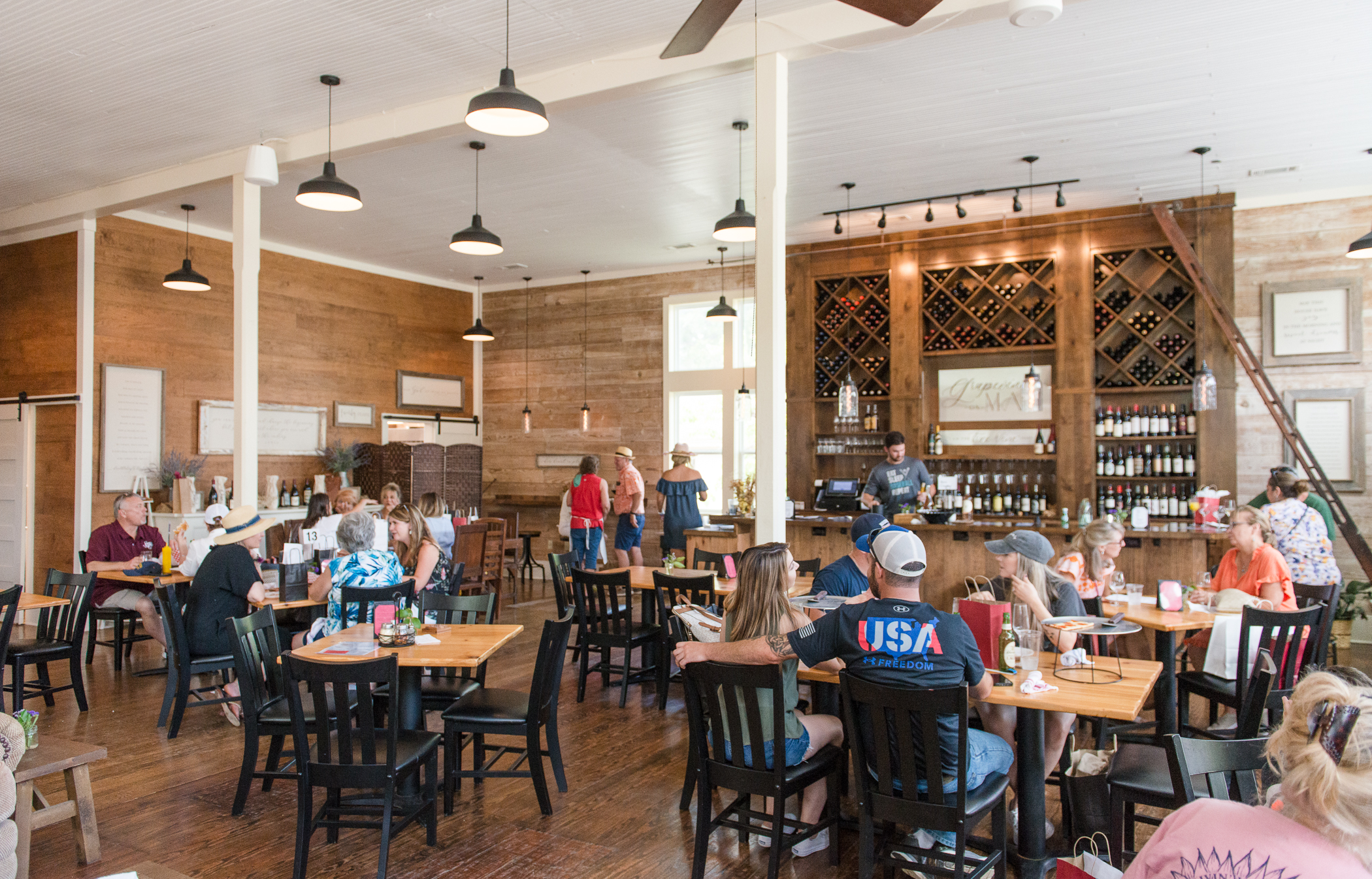 Many people sitting at tables talking to each other. In the back there is a bar with a server pouring drinks.