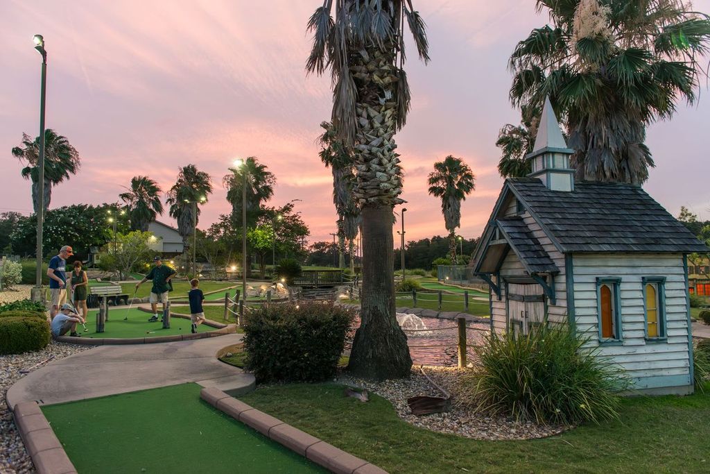 A miniature golf course at sunset. A family of 5 is playing one of the holes.