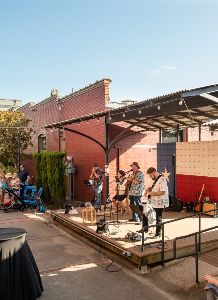 A local band performs on an outdoor stage in downtown Brenham, Texas. The musicians, dressed in casual summer attire, play guitars and harmonicas under a covered pavilion. A red brick building and a painted Texas flag backdrop set the scene, while families with strollers and onlookers gather nearby to enjoy the live music on a sunny day.