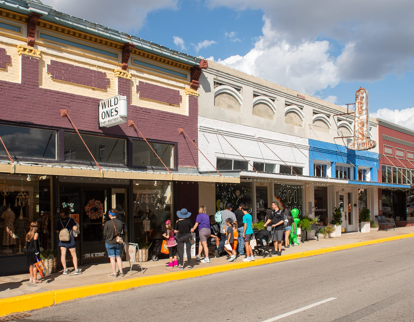 Families and children in costumes line up outside shops in downtown Brenham, Texas, during a trick-or-treat event. The storefronts are decorated for fall, and people carry candy bags while enjoying the festive atmosphere.