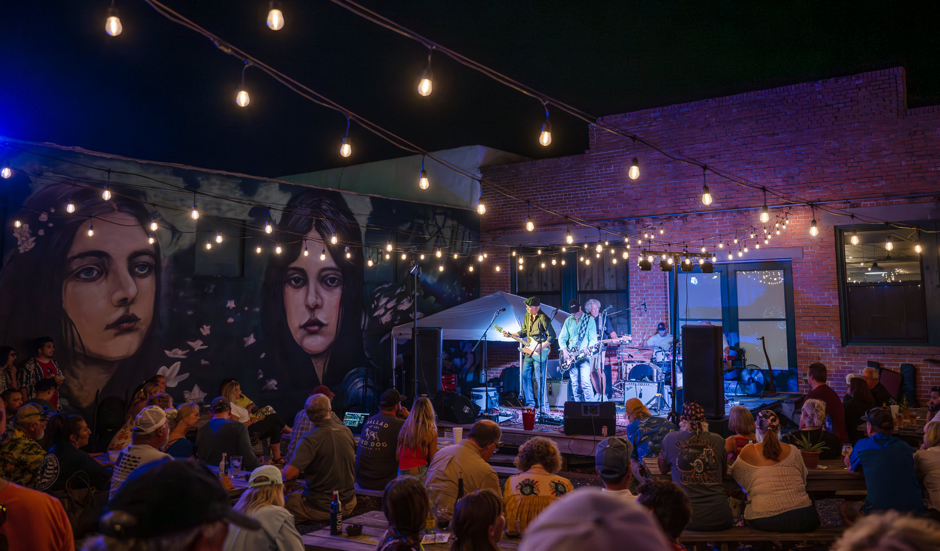 Outdoor concert at Home Sweet Farm Biergarten in Brenham, Texas, with a live band performing under string lights, a crowd of people seated at picnic tables, and a large mural of two women’s faces on the brick wall behind the stage.