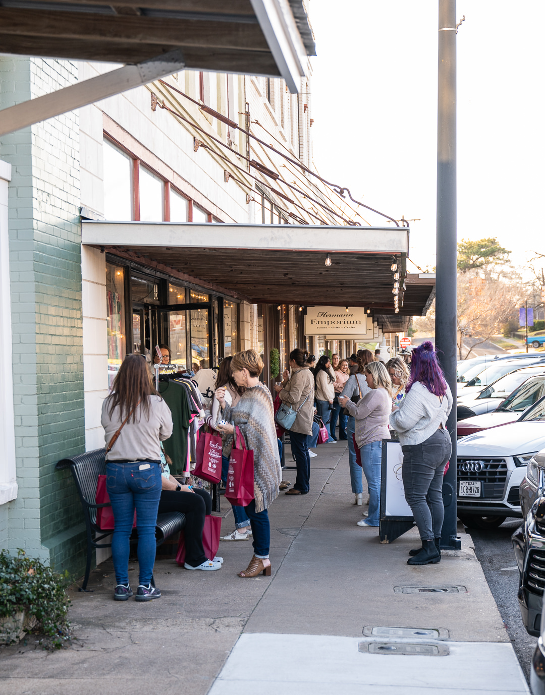 Uptown Swirl participants are gathered visiting outside of Anatomy clothing boutique and Hermann Emporium on a sunny day in Downtown Brenham.