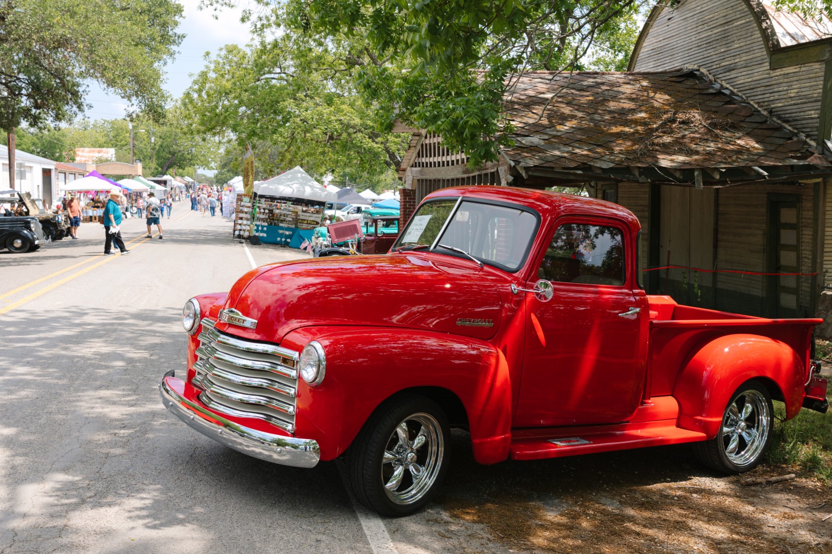 A bright red vintage Chevrolet pickup truck parked in front of an old wooden building during the Burton Cotton Gin Festival. In the background, festival-goers walk along a vendor-lined street with tents, crafts, and food stands under a sunny sky.