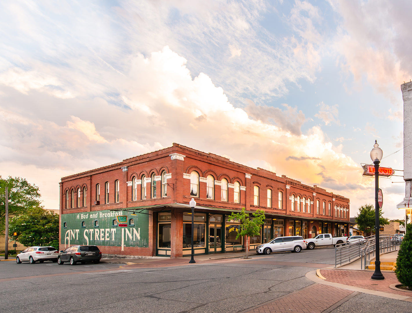 A wide view of the historic Ant Street Inn in Downtown Brenham, Texas, captured during golden hour with a glowing sunset sky. The two-story red brick building features arched windows, vintage signage reading “A Bed and Breakfast Ant Street Inn,” and a green painted mural along the side. Several cars are parked along the street, and a classic neon "Liquor" sign hangs from a nearby lamppost, adding to the old-town charm of the scene.