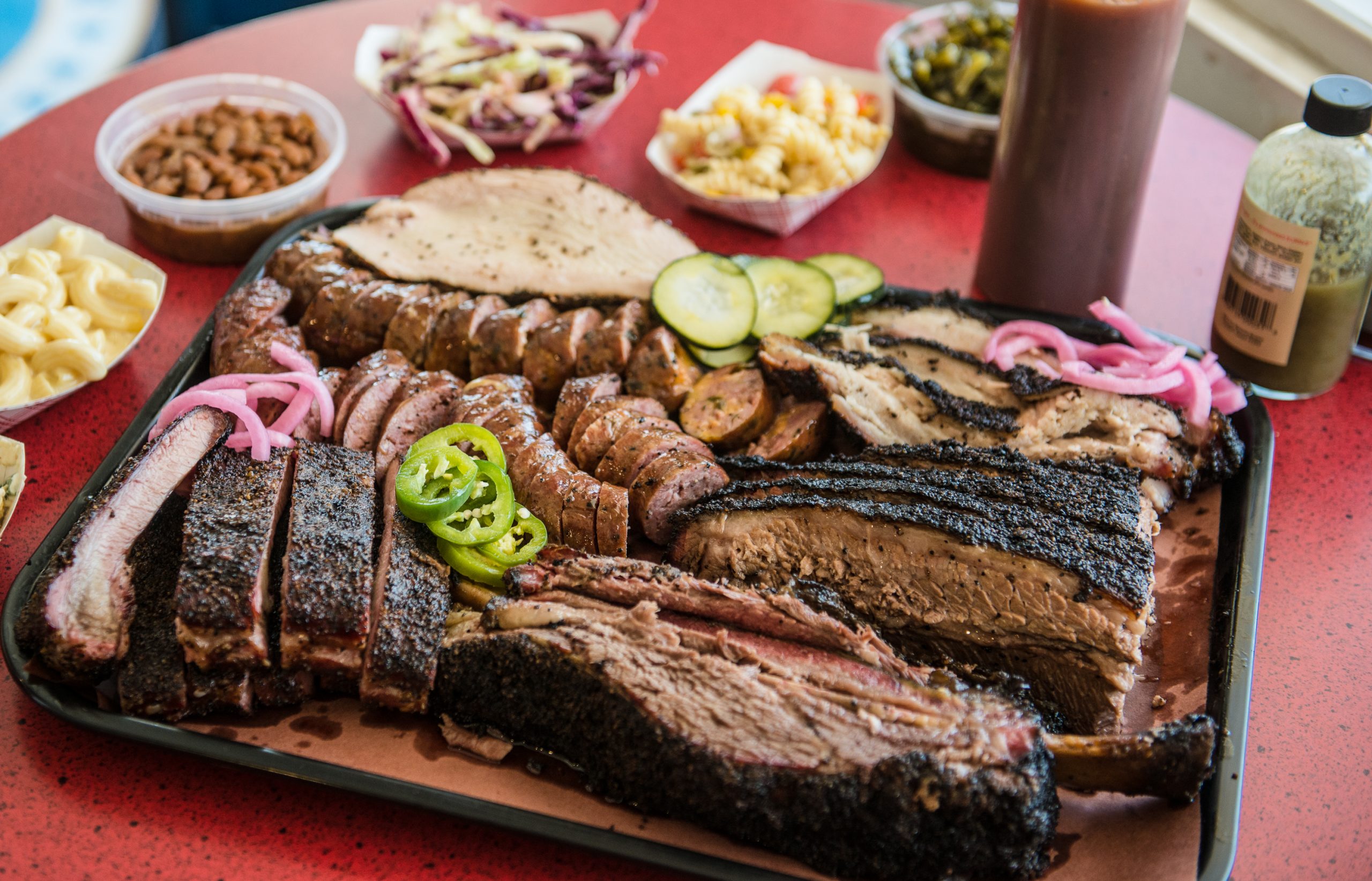 A hearty tray of Texas-style barbecue featuring an assortment of smoked meats including brisket, sausage, ribs, and beef ribs, garnished with sliced pickles, jalapeños, and pickled onions. Surrounding the tray on a red table are classic Southern sides such as baked beans, coleslaw, macaroni and cheese, and greens. Barbecue sauce and seasoning bottles are also visible, enhancing the authentic, mouthwatering spread.