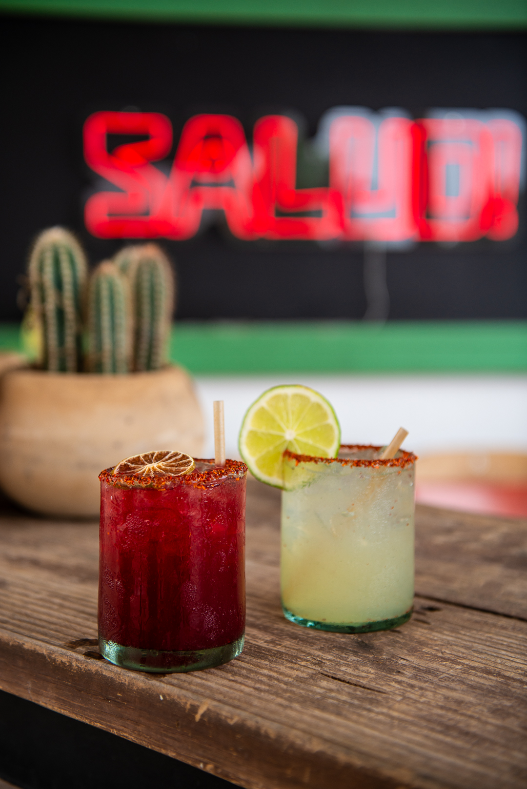 Two colorful cocktails sit on a rustic wooden table at Salud, a trendy bar known for its vibrant atmosphere. One drink is deep red with a chili salt rim and dried citrus garnish, while the other is a light yellow cocktail garnished with a lime wheel and chili salt rim. A potted cactus adds a southwestern touch, and the neon red “SALUD” sign glows in the blurred background, enhancing the fun and festive vibe.