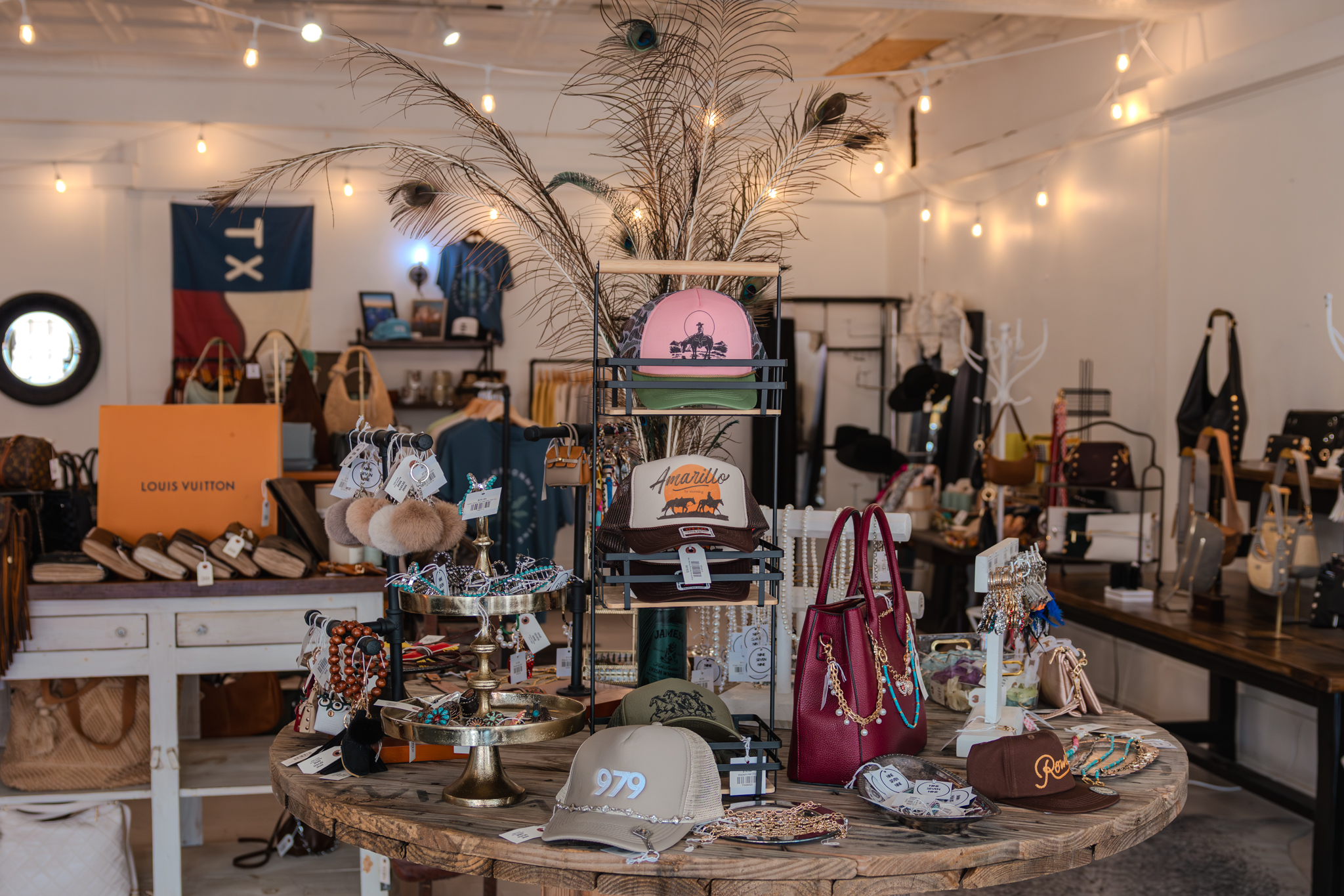 Interior of Nine Seven Nine, a boutique in Downtown Brenham, showcasing a display of trendy hats, jewelry, handbags, and accessories. A round wooden table features branded trucker hats, including one with “979” on the front, along with beaded bracelets, necklaces, and a red handbag. The background includes clothing racks, designer bags, and a Texas-themed flag hanging on the wall, creating a stylish and welcoming shopping atmosphere.