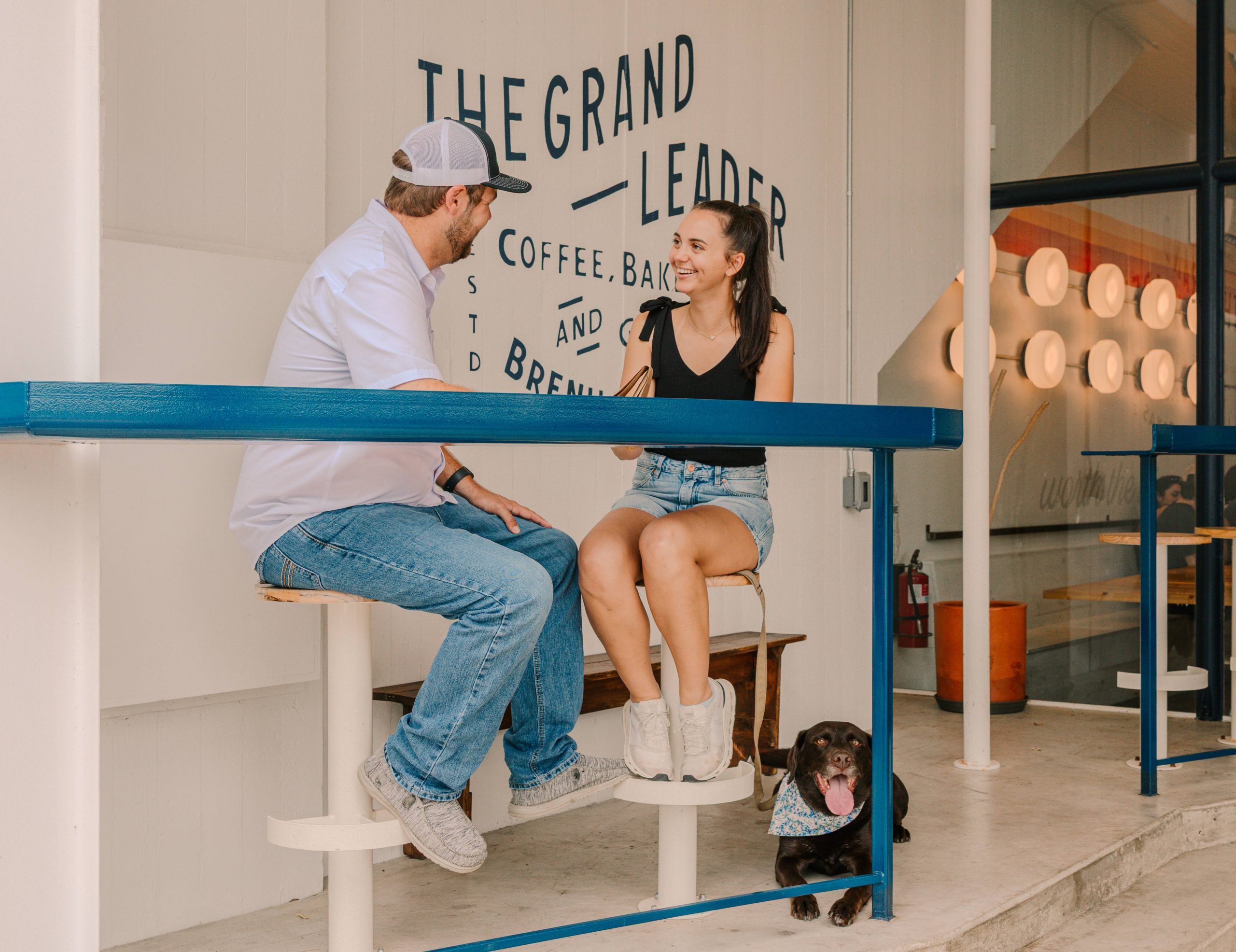 A man and woman sit at the outdoor counter of Mescalito Coffee in Downtown Brenham, smiling and enjoying their drinks. Their chocolate lab rests happily at their feet, panting and wearing a floral bandana. The café’s modern signage is visible behind them.