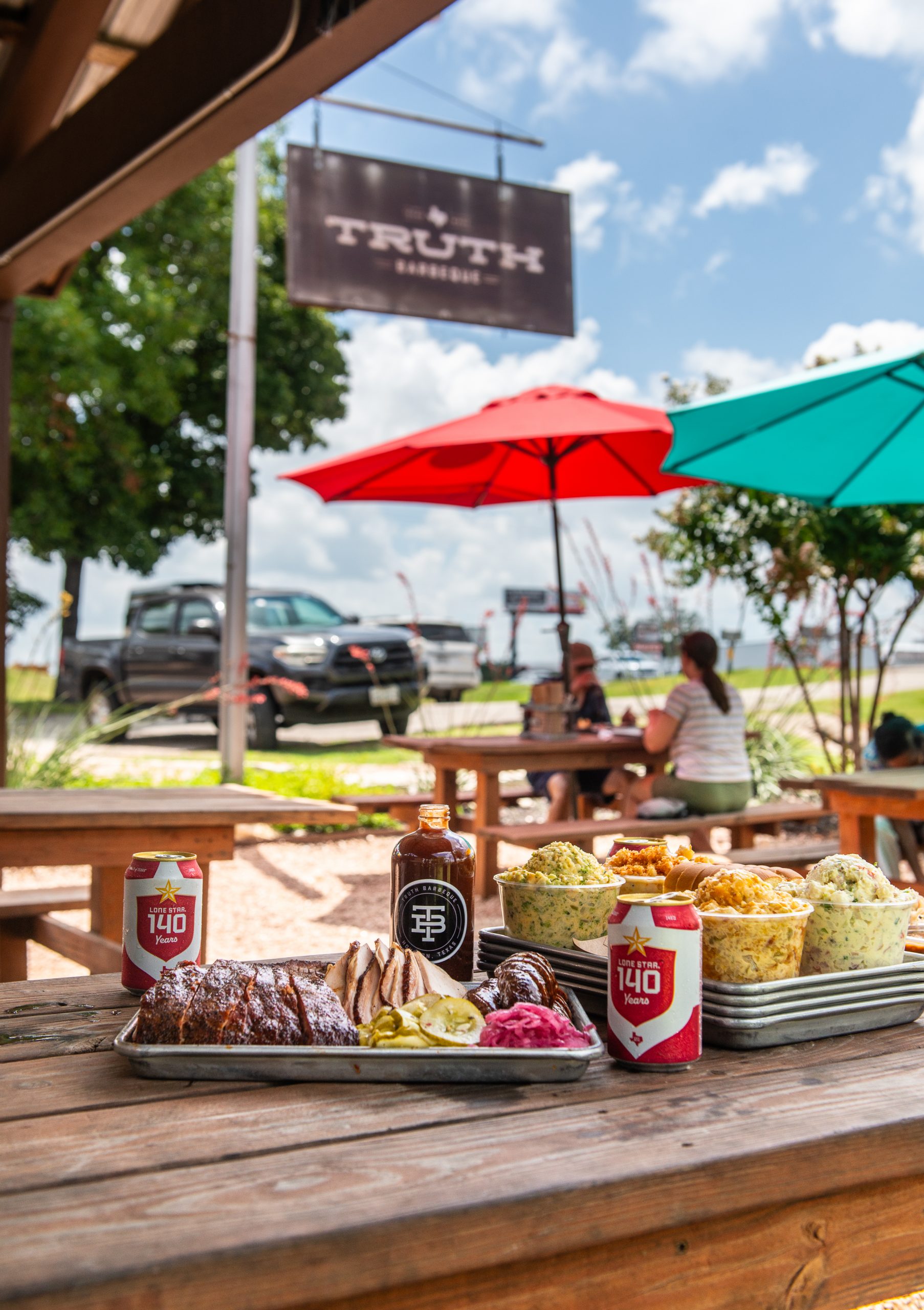 A picnic table at Truth BBQ showcases a mouthwatering Texas barbecue feast with trays of smoked meats, pickled vegetables, and an assortment of creamy side dishes like potato salad and coleslaw. Two cans of Lone Star beer and a bottle of house barbecue sauce are also on the table. In the background, diners sit under colorful red and teal umbrellas, and a rustic "Truth Barbeque" sign hangs above, set against a sunny sky with fluffy clouds. The outdoor setting features wooden benches, gravel ground, and nearby parked trucks, evoking a relaxed, classic Texas barbecue experience.
