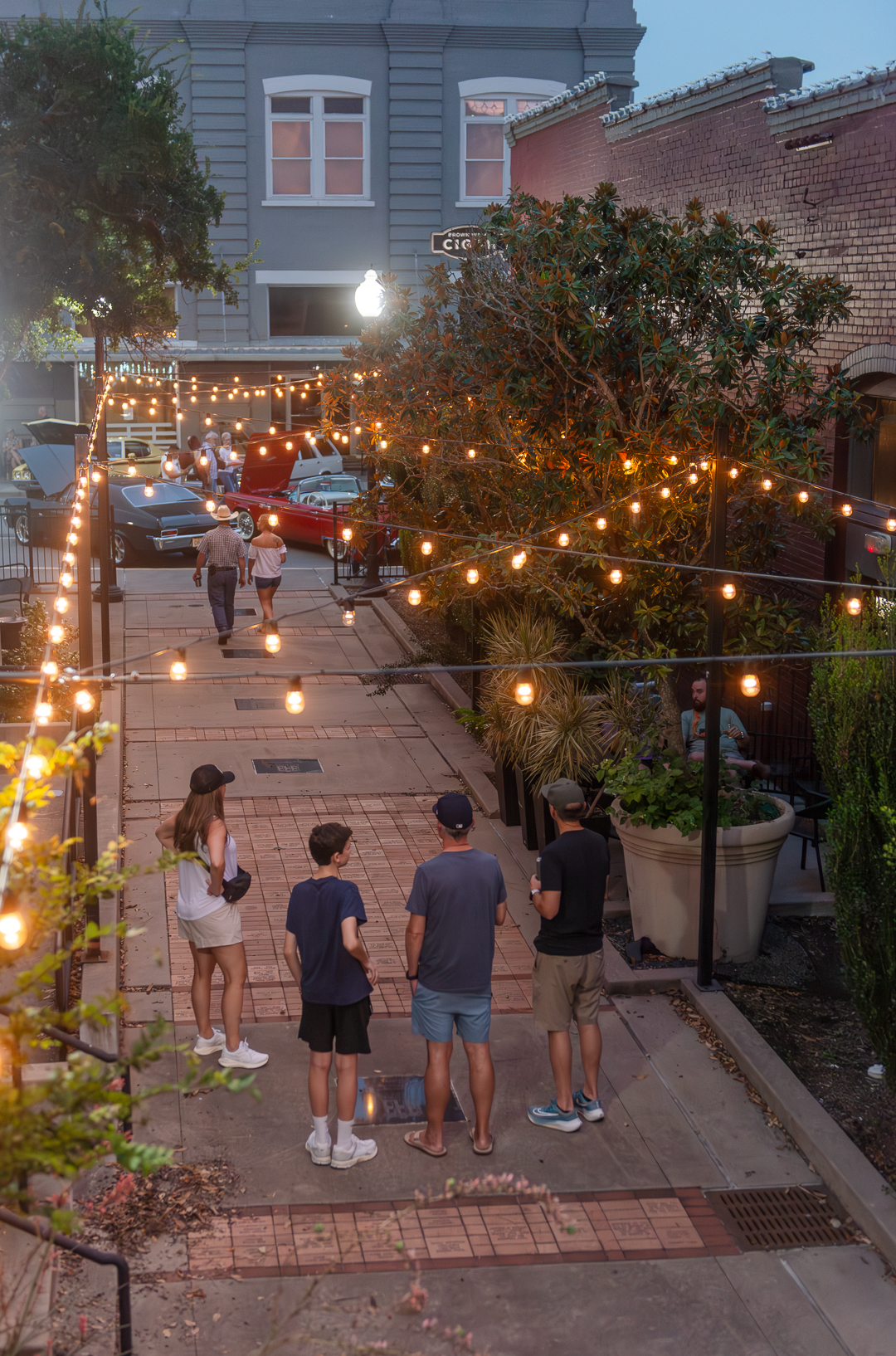 An evening scene in Alamo Alley in Downtown Brenham, Texas, softly lit by string lights suspended overhead. A group of four people stands in the foreground, taking in the view of a nearby classic car show, where vintage vehicles with open hoods are parked along the adjacent street. The alley is lined with trees, planters, and cozy seating areas, creating a warm and inviting atmosphere. The soft glow of the lights and the relaxed crowd evoke a charming, small-town summer night.