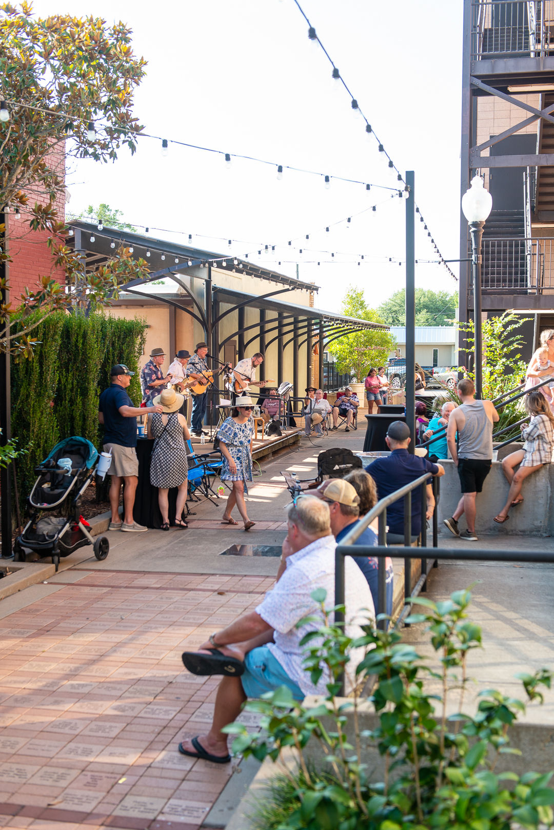 A lively outdoor concert takes place in Alamo Alley in Downtown Brenham, Texas. A local band performs on a small stage under a covered awning, surrounded by greenery and string lights. Audience members of all ages relax in folding chairs, lean against railings, or sit on low walls, enjoying the music. The scene is vibrant and casual, with families, couples, and friends gathered on a sunny summer afternoon. A stroller, coolers, and sun hats add to the laid-back, community feel of the event.