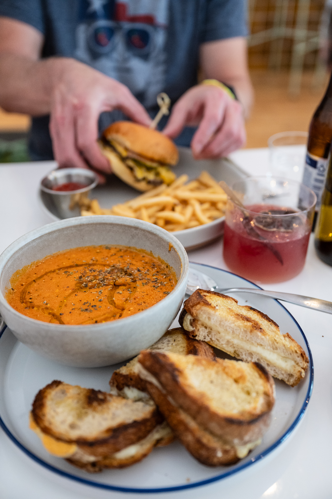 A cozy meal at Danny's, featuring a comforting bowl of tomato basil soup drizzled with olive oil and herbs, served alongside a plate of golden grilled cheese sandwiches. In the background, a person prepares to enjoy a cheeseburger and fries with a side of ketchup. The table also includes a pink cocktail with fresh herbs and a bottle of beer, capturing a relaxed, casual dining experience with elevated comfort food.