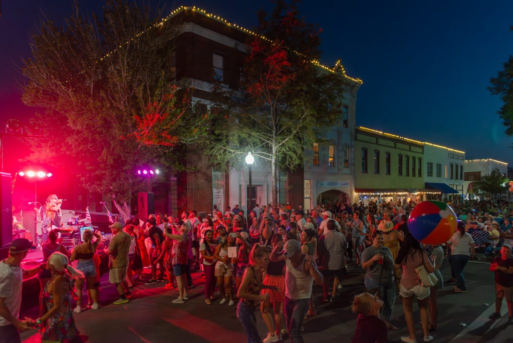 A vibrant nighttime street concert in downtown Brenham, Texas, featuring a lively crowd dancing and enjoying live music under colorful stage lights. The performer sings energetically on stage as red and purple lights illuminate the surrounding trees and buildings. Attendees, including families and groups of friends, fill the street, some dancing and others tossing a large beach ball. Historic buildings decorated with string lights provide a charming backdrop, adding to the festive and electric atmosphere of the summer evening event.