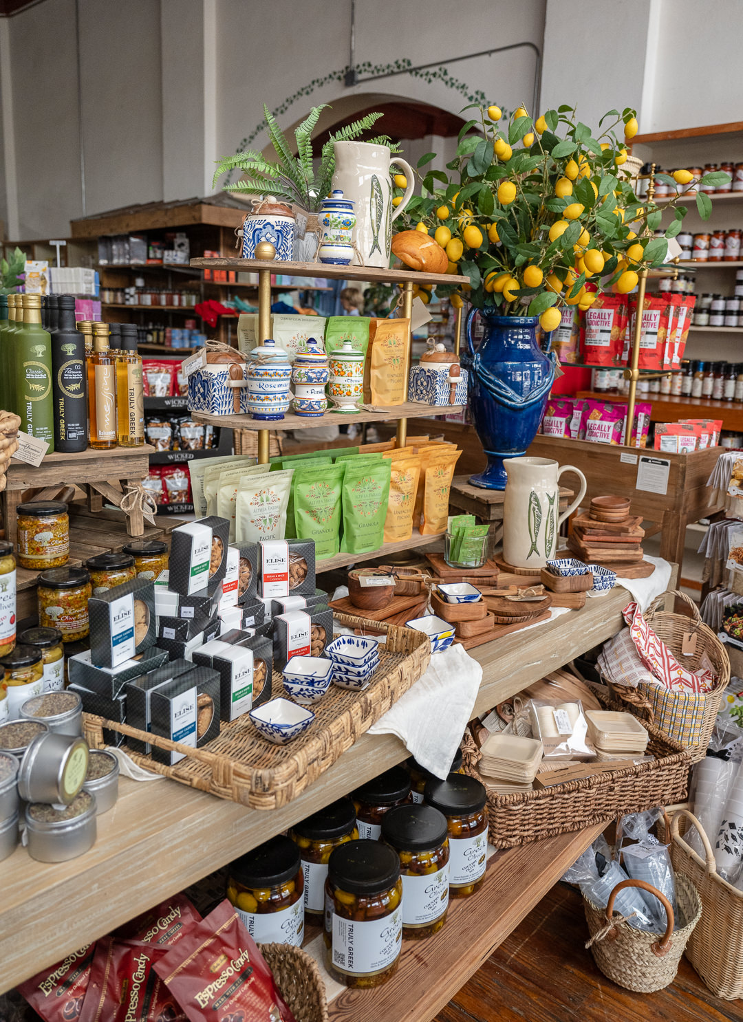 A colorful and carefully curated display at Hermann Emporium, showcasing gourmet foods, artisanal kitchenware, and home decor. Shelves are filled with olive oils, spice blends, herbal teas, and jars of truffle goods. Decorative ceramic jars, wooden bowls, and hand-painted pottery are artfully arranged alongside woven baskets and linen textiles. A vibrant blue vase filled with faux lemons adds a cheerful touch, enhancing the warm and inviting ambiance of this specialty shop.