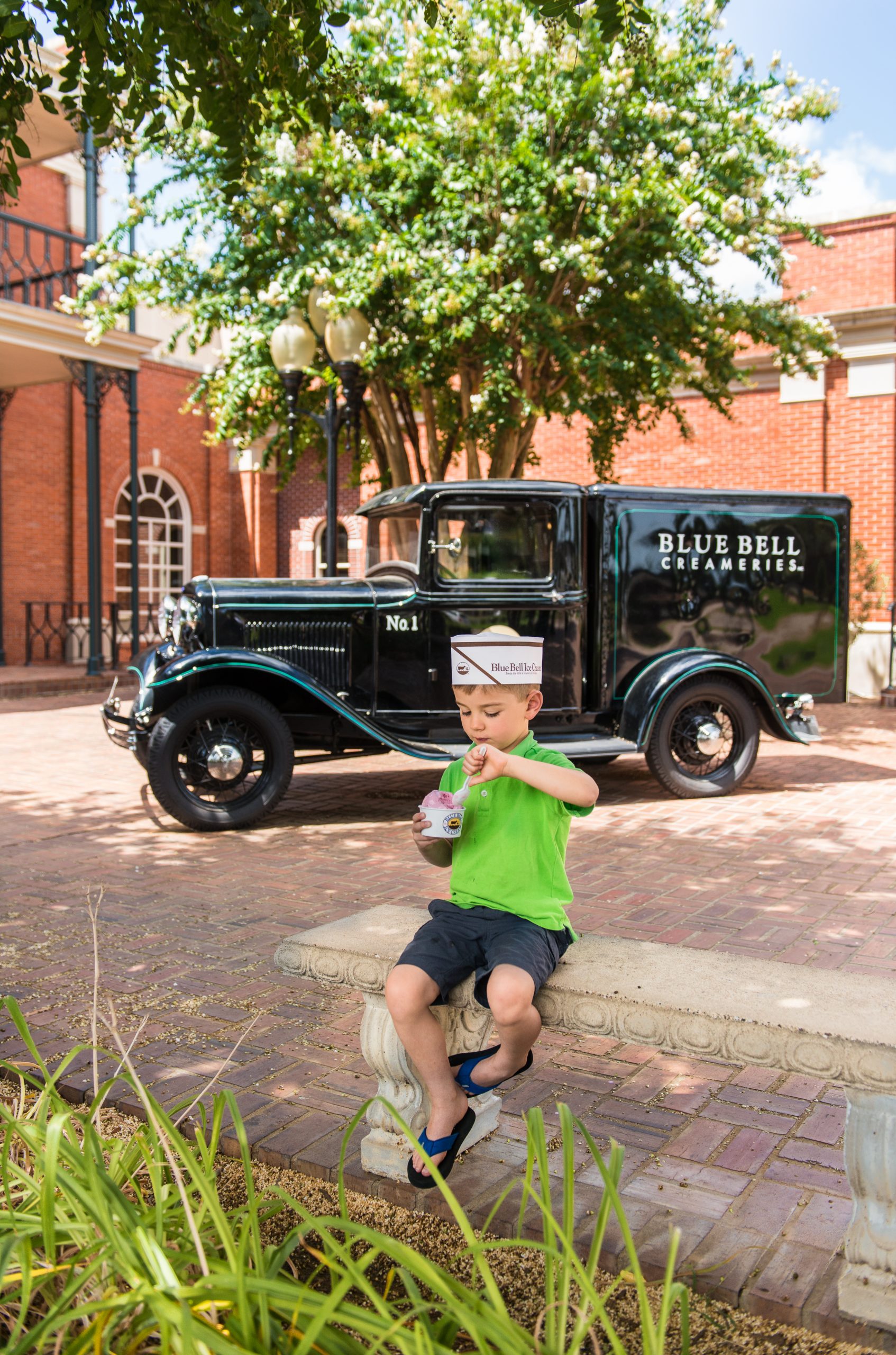 A young boy wearing a green shirt, navy shorts, flip-flops, and a paper Blue Bell Ice Cream hat sits on a decorative stone bench enjoying a cup of ice cream. Behind him is a vintage black Blue Bell Creameries delivery truck parked on a brick courtyard. The background features red brick buildings, a blooming crepe myrtle tree, and old-fashioned street lamps, creating a nostalgic and charming atmosphere.