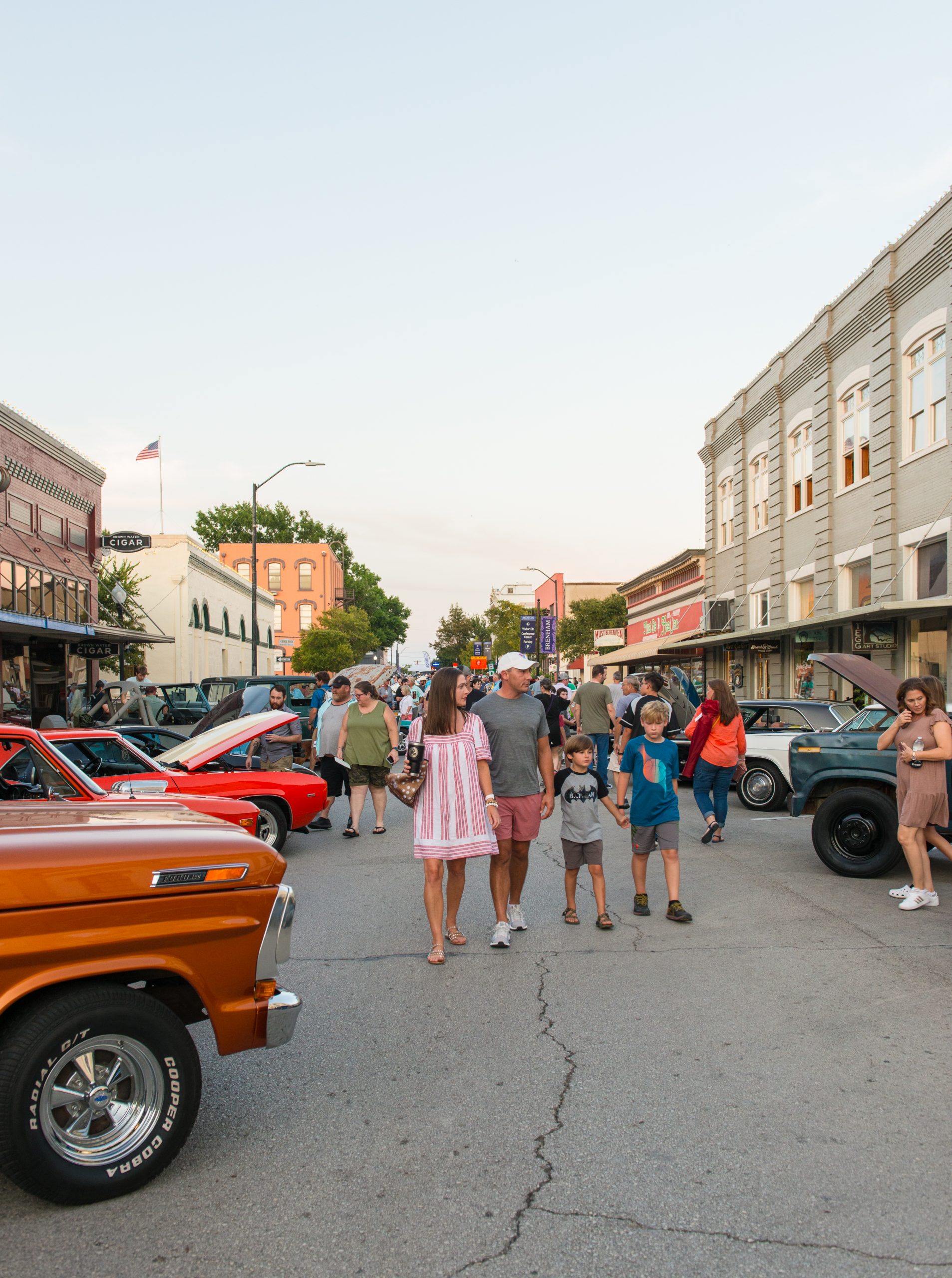Families and visitors stroll down a blocked-off street in Downtown Brenham, Texas during a classic car show. Vintage cars with open hoods line both sides of the road, while people admire the vehicles and enjoy the evening atmosphere. Historic buildings and local shops frame the scene, and the sky is clear with a soft glow from the setting sun. A couple holds hands with their two young sons in the foreground, adding to the family-friendly, small-town charm of the event.