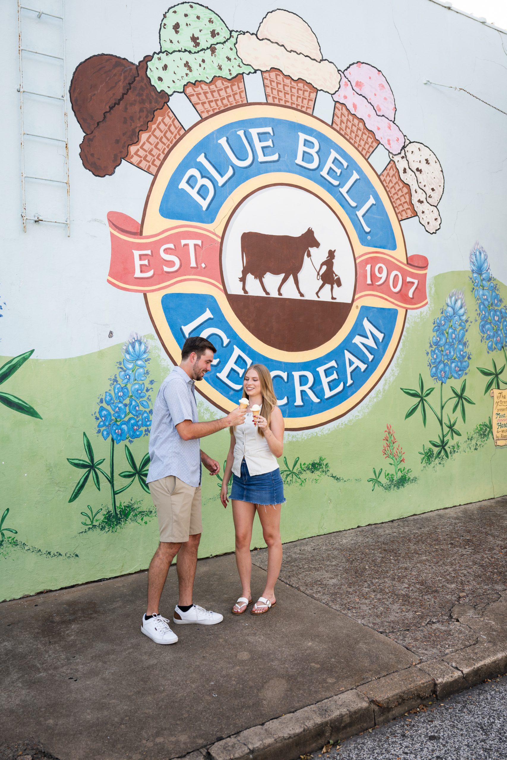 A smiling couple enjoys Blue Bell Ice Cream in front of the iconic Blue Bell mural in Brenham, Texas, featuring colorful ice cream cones and the classic logo with a cow and child. The scene is cheerful and summery, capturing the charm of the Ice Cream Capital of Texas.