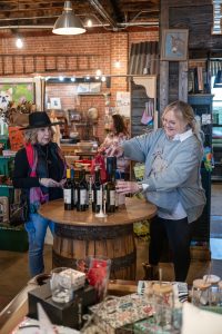 Two women participate in a wine tasting at Uptown Swirl. One woman, wearing glasses and a light blue sweatshirt, pours wine into a cup at a round barrel table lined with bottles. Another woman, dressed in a black hat, vest, and colorful scarf, watches as she prepares to taste. The cozy shop has rustic brick walls, warm lighting, and shelves of local goods in the background.