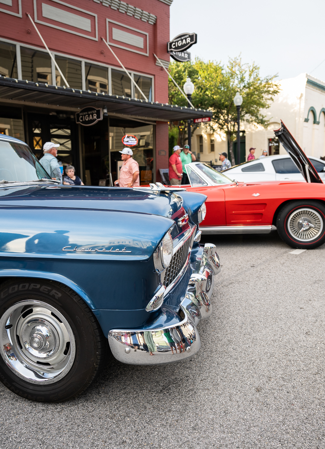 Classic cars on display during Downtown Brenham’s Hot Nights, Cool Tunes event. A shiny blue vintage Chevrolet is parked in front of a red convertible with its hood open, while people gather on the sidewalk near a cigar shop. The scene captures the lively atmosphere of the summer event, where music, community, and classic cars take over the streets.