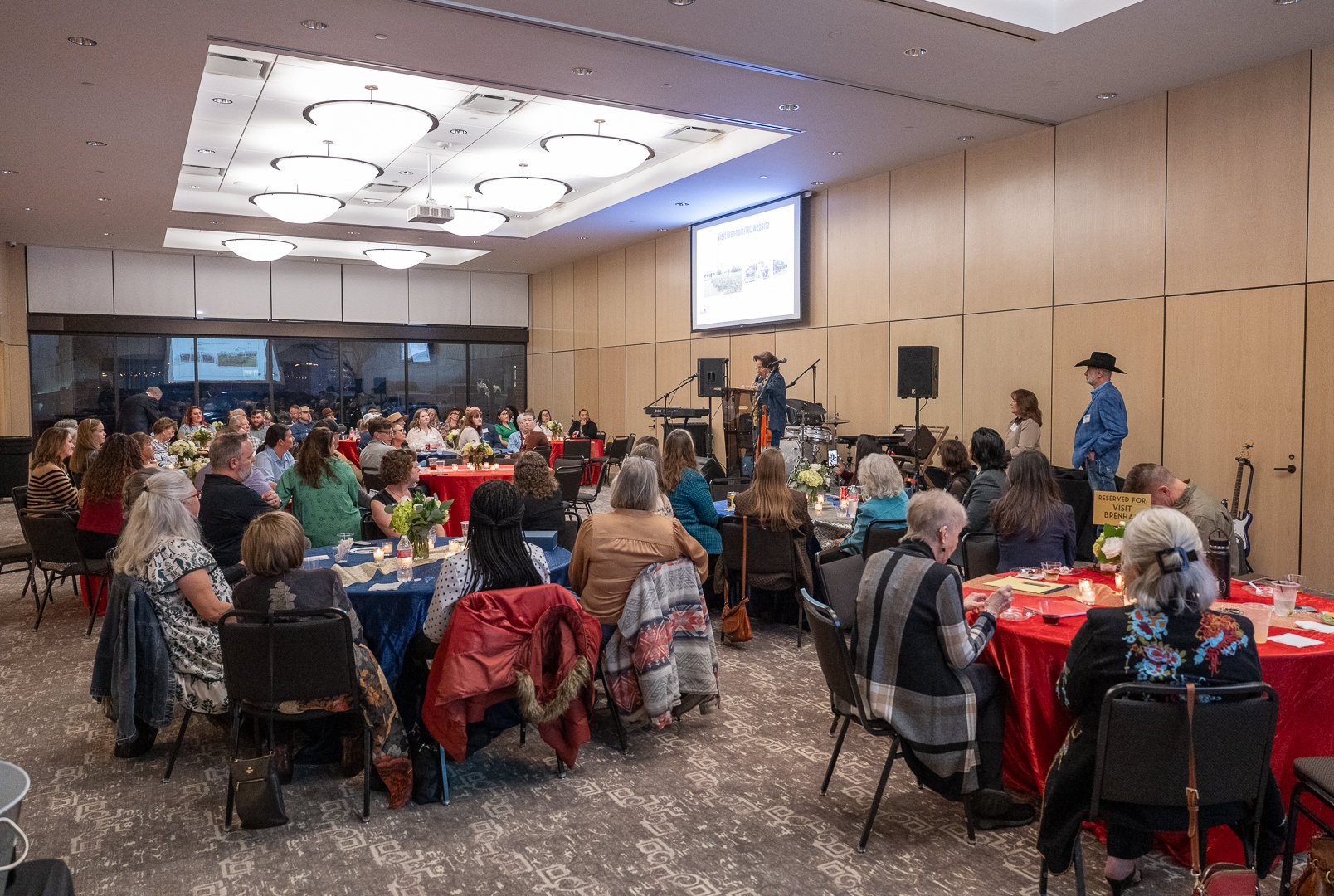 Guests gather at round tables for the Tourism Partner Awards at The Barnhill Center in Brenham, Texas, as speakers and musicians stand on stage beneath a projected presentation.
